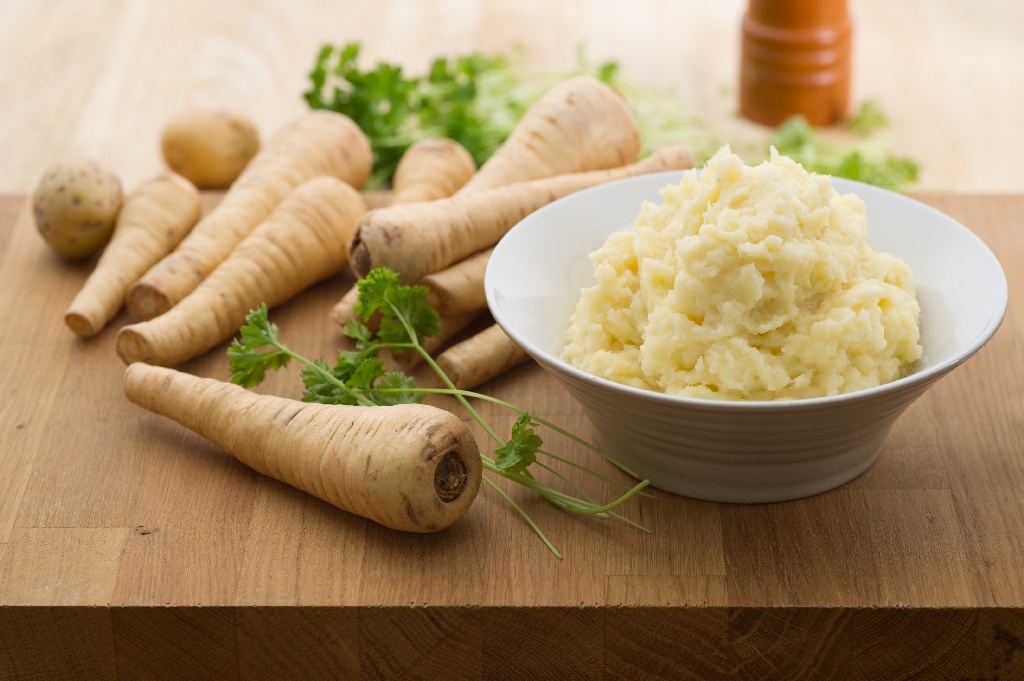 A bowl of parsnip and potato mash, on a cutting board next to a number of parsnips, and parsley.