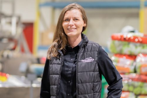 Image of a woman in a black Gwillimdale Farms vest. She is looking at the camera and smiling. The background is out of focus, but stacks of bags of produce and metal shelving are visible.