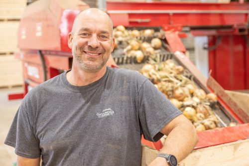 Image of a man wearing a grey Gwillimdale Farms T-shirt. He is smiling for the camera, his elbow resting on a wooden crate. Above the crate is a inclined conveyor, carrying freshly harvested onions.