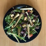 A salad made with parsnips, and a a variety of greens, in a black dish, photographed on a wood table from above.