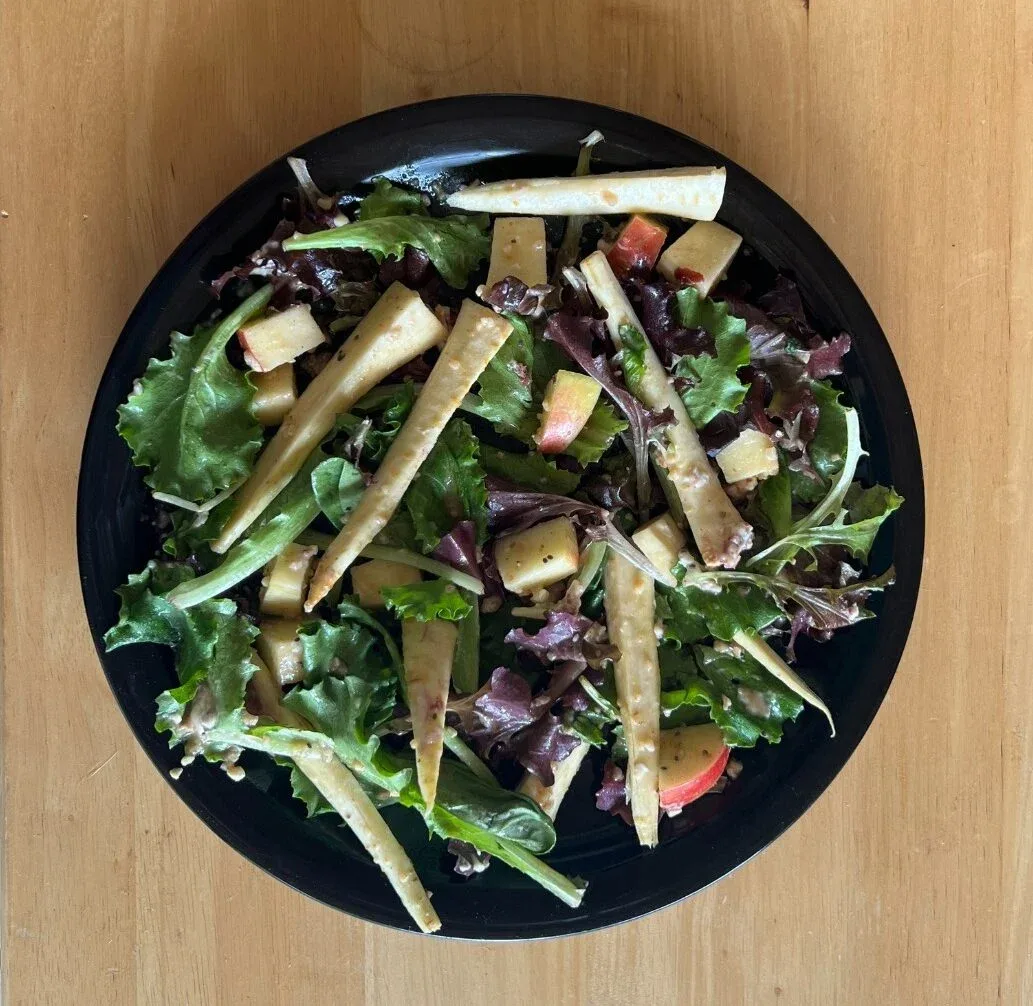 A salad made with parsnips, and a a variety of greens, in a black dish, photographed on a wood table from above.