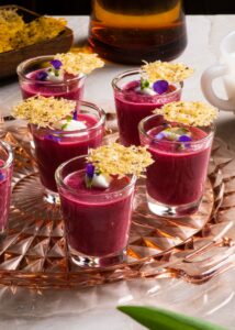 Five shot glasses filled with a thick, vibrant fuschia-coloured liquid, and topped with violet pedals, micro-greens, and a parmesan crisp.