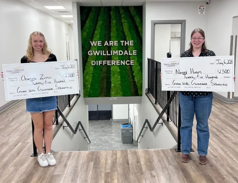 Two young women standing in front of a wall mural depicting art of rows of deep green crops, behind the words "We Are the Gwillimdale Difference" in white. They are both smiling, while holding up very large cheques. The cheques have the Gwilimdale Farms logo in the top left corner, the date "July 6, 2023" in the top right. Center right is the label "Pay to the order of" followed by the students' names, and the quantity "$2500.00". Across the bottom of the cheque is a label, "Memo:" followed by the words "Grow with Gwillimdale Scholarship"