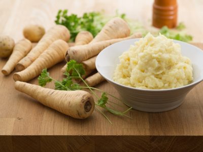 A bowl of parsnip and potato mash, on a cutting board next to a number of parsnips, and parsley.